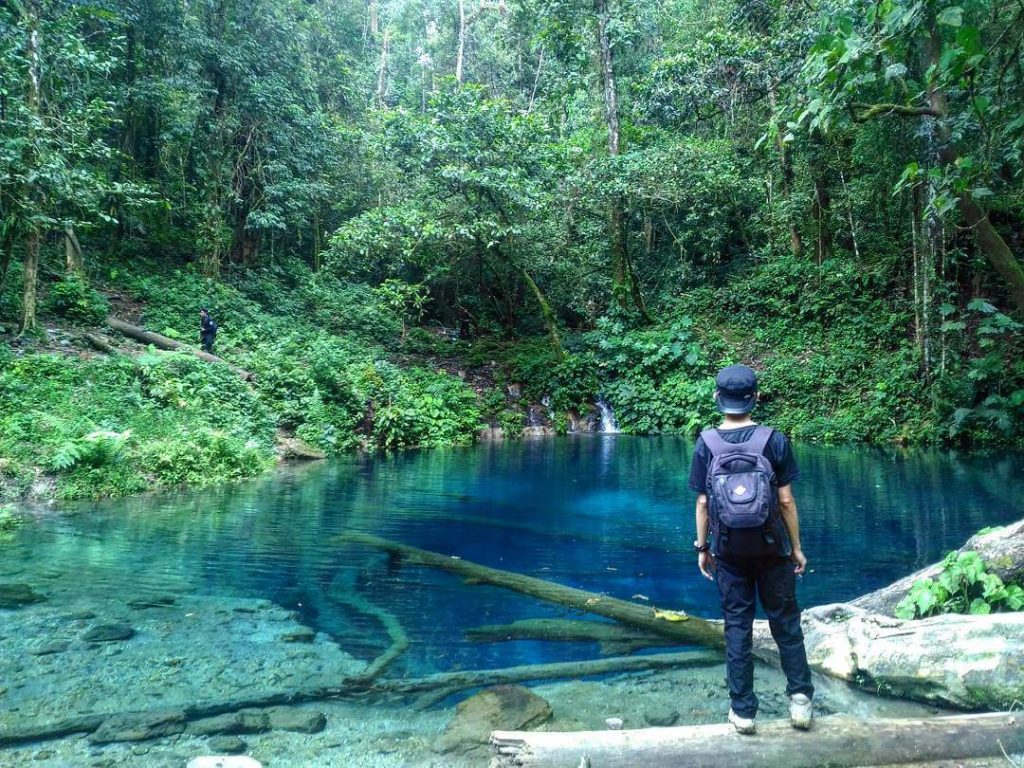 Seorang pria memakai topi, berdiri di pinggir Danau Kaco yang airnya terlihat biru jernih.